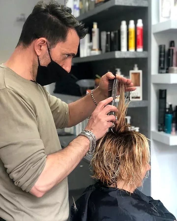 Matteo taglia i capelli di una cliente, con gli scaffali dei prodotti alle spalle
